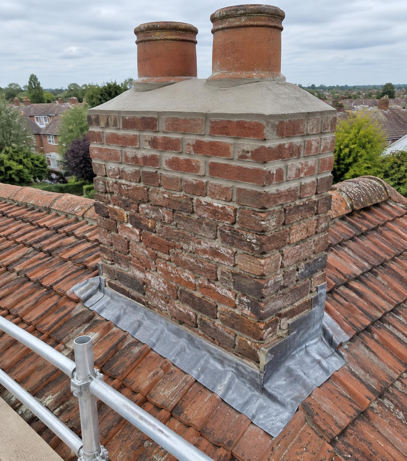 Repointed brick chimney stack with fresh flaunching around terracotta pots and new lead apron, viewed from scaffolding